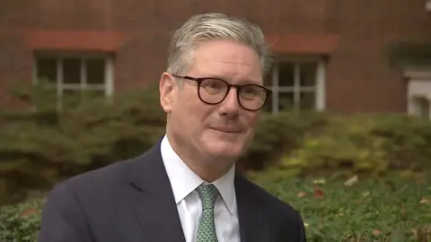 Prime Minister, Sir Keir Starmer, looks slightly off to the right of the camera while talking to Peter Henley, he is wearing slightly rounded black framed glasses, and green patterned tie with a white shirt and dark grey blazer and stands infront of a red brick building with white windows and greenery