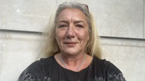 Melissa Moore stands outside her office against a wall, wearing a dark top, silver necklace, with glasses holding her blonde hair in place