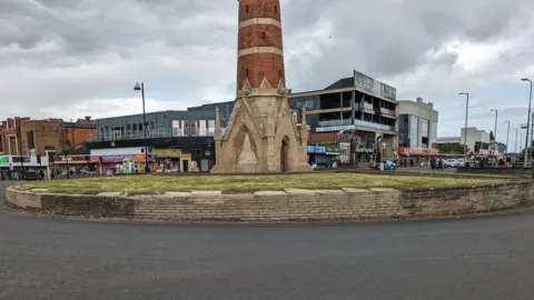 Lincolnshire County Council A landscape street view of a circular raised lawn surrounded by a low wall on a tarmac road. The wall is discoloured on the left and right with the yellow/beige stone bricks missing. In the middle is a narrow red brick building shaped like a tower with the base made of stone and featuring gothic edifices. In the background are retail units and commercial buildings