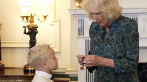PA Rupert Williams standing in front of Queen Camilla. Rupert wears a white short sleeved shirt, a beige waistcoat and a black bow tie. Queen Camilla wears a patterned green, blue and orange dress. The Queen holds his medal. 