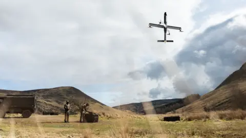 STARK Two people, bottom left, stand next to equipment, which is flying a drone that is captured in the sky to the right of the photo. They are standing in a hilly landscape with trimmed grass