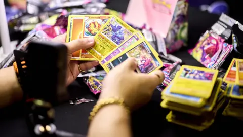 London Card Show Two hands holding multi-coloured Pokemon cards in front of a camera.