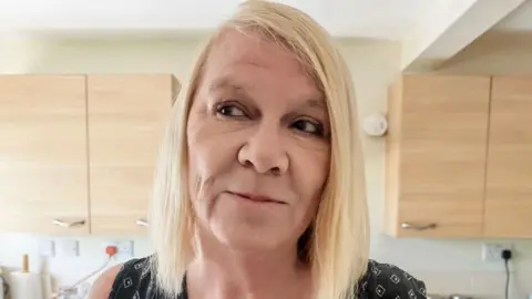 Jaqueline Watkins standing in front of cupboards. She is looking off to the side of the picture. She has shoulder-length blonde hair, with a side fringe.