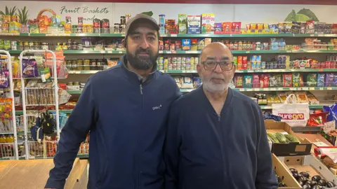 Two men, one on the right in his 60s, the one on the left in his 40s, standing in front of a shop