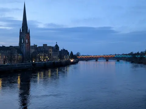 Ian Fuller A view looking across water, with a lit up bridge in the distance. Nearer to the camera is a city centre, with a church and steeple prominent. 
