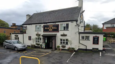 Google Maps Street view of the exterior of a cream-coloured pub with black detailing. The gold lettering on the building reads 'The Black Bull Inn'. There is a tarmac-surfaced car park in the foreground.