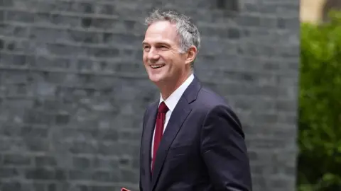 PA Media A man with grey hair in a dark suit, white shirt and maroon tie is walking past a brick wall and hedge.