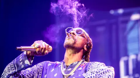 EPA Snoop Dogg در حالی که میکروفون در دست دارد و روی صحنه اجرا می کند سیگار می کشد