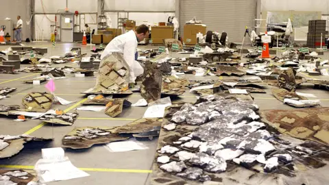 The wreckage of NASA's Space Shuttle Columbia is laid out on the floor of a large coat rack. A man in a white coat leans down and examines some of them. There are tiles and pieces of white foam - some of the material is charred.