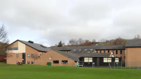 A general view of Crickhowell High School, a group of red brick buildings with a grass bank in front of it and a metal fence to the right. Trees can be seen in the horizon. 