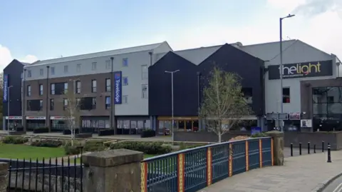 A number of connected buildings, including a Travelodge and The Light cinema, are set back from the grassy bank of a river. In the foreground, a bridge crosses the river.