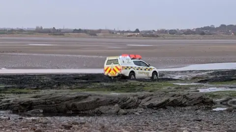 Wirral Coastguard A white van with blue and white insignia and the word 'Coastguard' along the side, parked on rocky patch on a beach. 