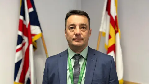 A man in a suit standing in front of a union flag and a Guernsey flag.