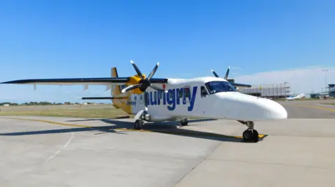 A yellow and white Aurigny Dornier aeroplane sits on an airport taxi way. It is quite small, capable of holding about 20 people, and has two propellors.