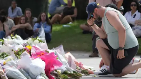 A man kneels in front of a large pile of flowers placed at Bondi Beach. Sitting on the grass around the pavilion, people are gathering.