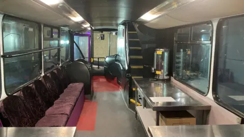 Interior of the lower deck of a double-decker bus, which has been converted to have a row of seats on the left, and a table with a kettle on the right.