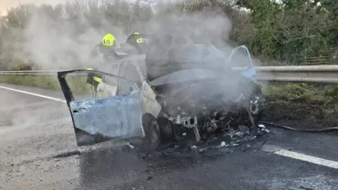 The picture shows a badly burned car on the side of a road. The front of the vehicle is almost completely destroyed, with the engine area exposed and charred. Thick smoke is rising from the wreckage, suggesting the fire has only recently been put out. The car doors are open, and the paintwork is scorched and peeling. Two firefighters wearing high-visibility helmets and protective gear are standing behind the car, partially obscured by the smoke.