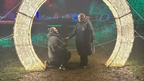Contributed A woman is down on one knee proposing to another woman. They stand in the middle of a large, circular light display. They are both wearing black coats and black trousers.