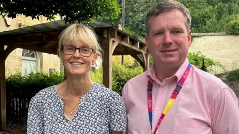A woman and a man smile at the camera outside the BBC Cornwall office. The woman has blonde hair and wears a summer dress, The man has dark hair and wears a pick shirt and an NHS lanyard.