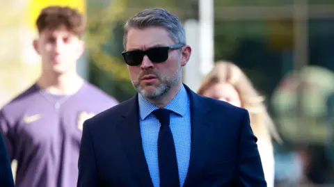 PA Media Adam Smyth wears a dark suit, a blue checkered collared shirt, a navy tie and black sunglasses. He has short greying hair and has a beard. His expression is neutral. Behind him are two people, but they are blurred.