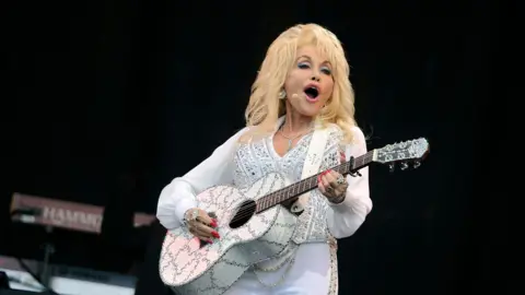 Country singer Dolly Parton, wearing a rhinestone-encrusted white suit, and playing a bejewelled guitar, on stage.