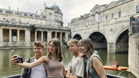 A group of young tourists taking a selfie in front of Bath's waterways and tunnels on an overcast day.