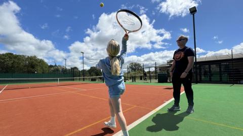 A young girl serves in Tennis while her coach watches on
