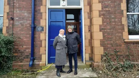 A woman wearing a grey coat and a man wearing a black jacket, stood in front of blue doors at the entrance of a red‑brick school building.