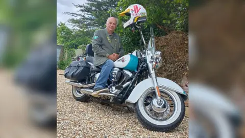 Paul sitting astride a shiny chrome classic motorbike with his helmet perched on the handlebars. He's wearig a grey casual jacket and jeans and is on a gravel drive