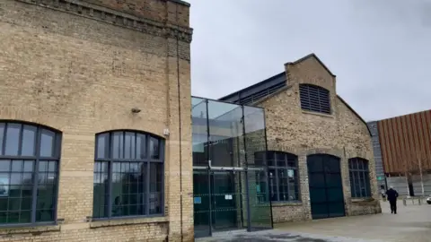 Sand Martin House in Peterborough. It is a listed former Victorian brick railway building containing two locomotive sheds transformed into offices. In between the buildings is is a modern black framed glass entrance and atrium, linking them. On the right a man in black can be seen walking away from the building. 