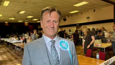 A man in a grey suit standing in an election hall. He has a light blue and white badge on he chest,