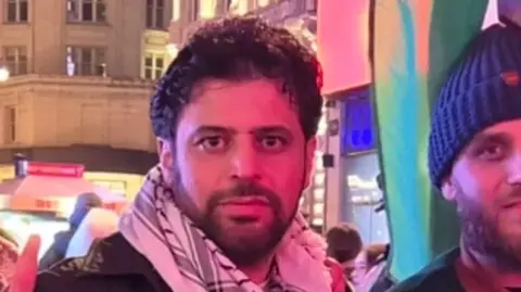 Abdulaziz Almashi Two men, Abdulaziz Almashi is pictured at night in front of buildings and people in the background 