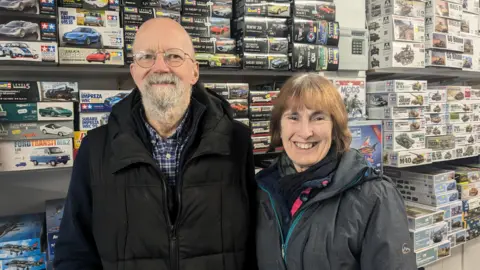 Paul, a white man with round silver glasses and a black coat with his wife Sue, who is white with short brown hair and a grey and blue coat. 