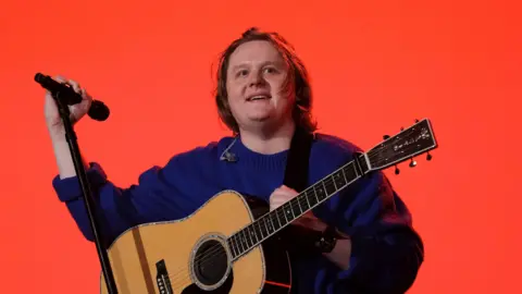 Getty Images Lewis Capaldi, a man with shoulder length hair, is pictured on stage at the Isle of Wight Festival. He is stoog against an orange backdrop wearing a blue jumper and a guitar on a strap. He is holding a microphone on a stand in his right hand.