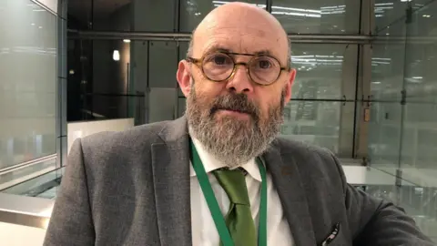 Vikki Irwin/BBC Andrew Stringer is pictured in an office with glass windows behind him. He is wearing a grey jacket and green tie.