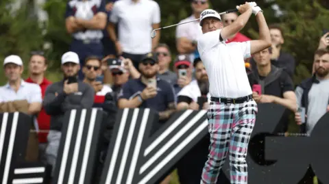 Ian Poulter in the middle of a follow-through after hitting a shot during a professional tournament. In the background are large black signs featuring bold white striped graphics.
