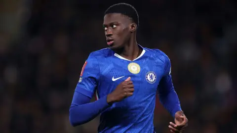 Mamdou Sarr visible from waist up running on the pitch against Hull City. He is wearing a blue Chelsea home jersey with matching blue undersleeves and is turning his head to look to his right