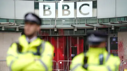 PA Media Image showing the paint splattered across the three revolving doors at the entrance to New Broadcasting House, while two out-of-focus uniformed police officers stand side by side 