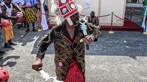 AFP via Getty Images A traditional dancer wearing an elaborate costume and headdress performs in front of a large wooden crate, while a crowd gathers around the area.