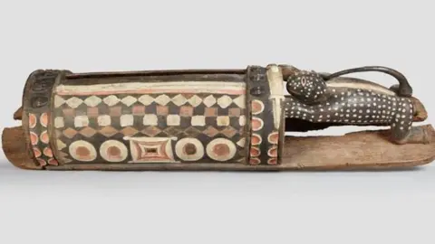 Ministry of Culture and Francophonie A large, ornately carved wooden talking drum, featuring geometric patterns and sculpted figures. 