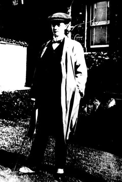 Mills Archive Trust A very over-exposed photo in black and white of a man wearing a flat cap, bow tie, long trenchcoat and shoes covered with white spats. He is leaning on a walking stick, despite not appearing to be very old.