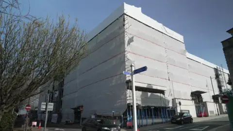 Evolution Cove is a large and tall building on the corner of two roads. It is covered in protective wrapping. There is a car parked on the pavement outside it and there are also traffic cones and warning signage on the pavement.