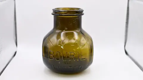 ODAIT A historic jar in a white display cabinet. It is brown.