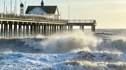 BBC Weather Watchers / Zingaro Blue Waves crash on the beach in Southwold, Suffolk; a pier can be seen on the left and the sky above is cloudy.