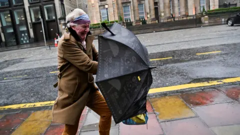 Reuters A woman struggles with an umbrella on George Street in Edinburgh. She is wearing a long brown coat with brown trousers and a multi-coloured head band. She is holding a black umbrella and grimacing against the wind.