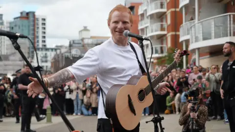 Shaun Whitmore/BBC Ed Sheeran performing at Ipswich Waterfront. He is performing to crowds of hundreds of people with a microphone and guitar. He is wearing a white T-shirt and is looking at the camera and smiling.