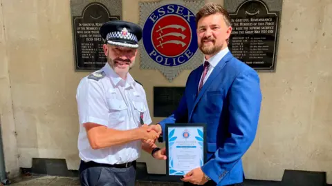 Essex Police Glen Pavelin is wearing his police uniform and is shaking Remblance's hand. Remblance is dressed in a blue shirt and navy tie and is holding the certificate as the award is presented.