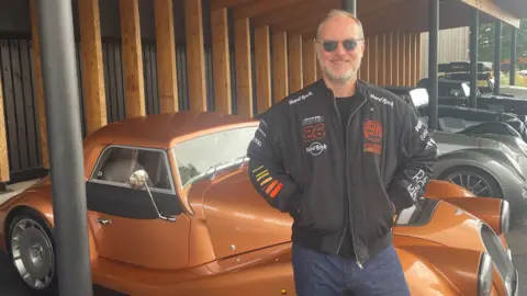 BBC A man in a black jacket and blue jeans stands alongside a row of Morgan sports cars. He is wearing sunglasses and has grey hair and a grey beard. 