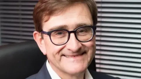 A man with glasses and brown hair sat in an office chair smiling into the camera.
