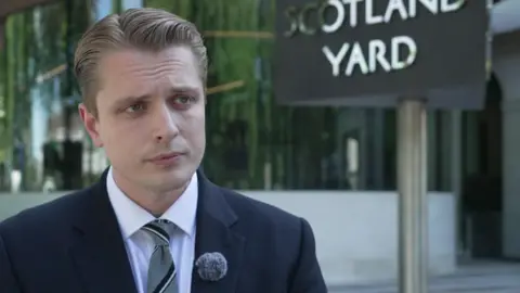Detective Superintendent Dan Thompson is wearing a dark coloured suit and a grey, striped tie, with a microphone attached to his lapel. He is standing outside New Scotland Yard. 
